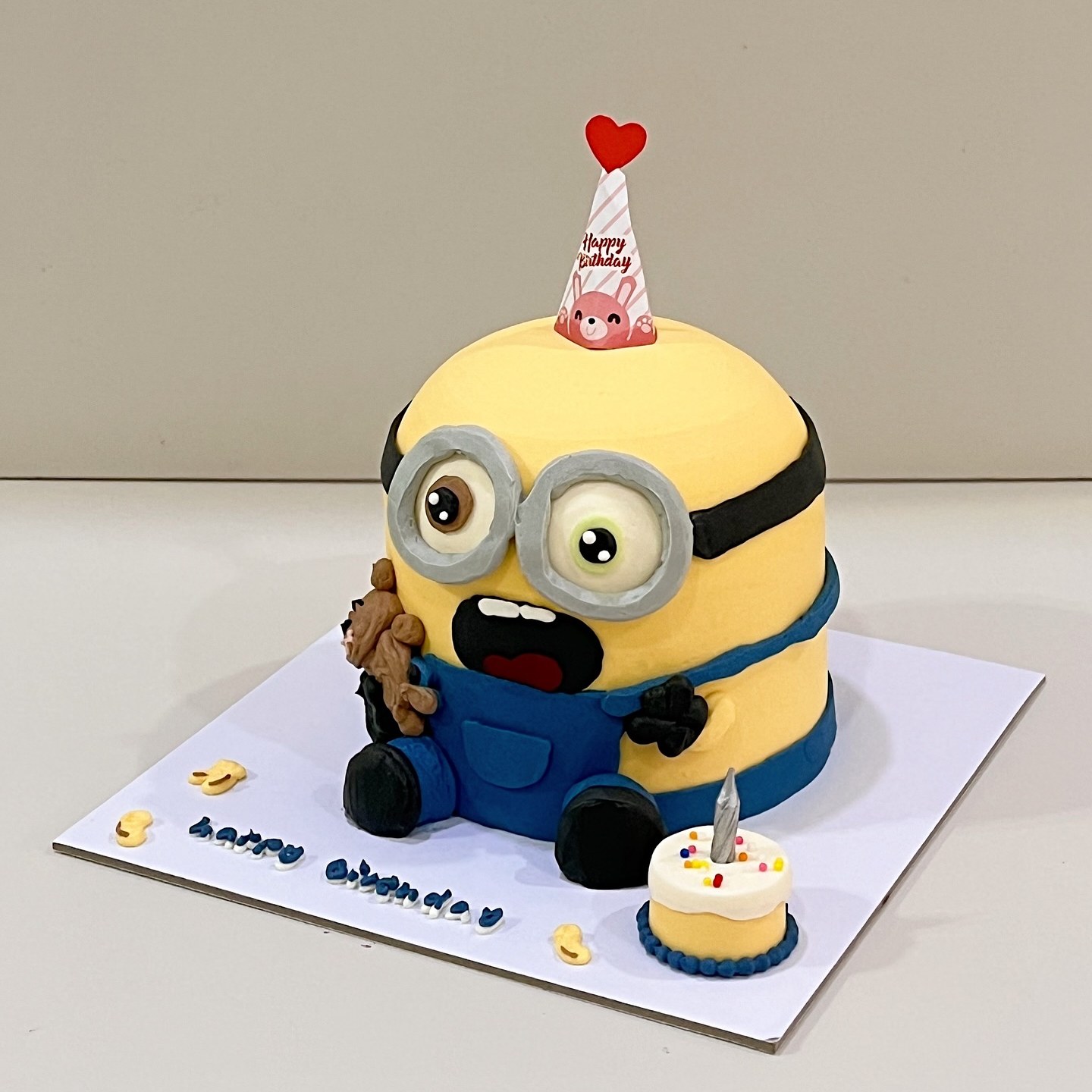 Goggle-eyed yellow character cake in blue overalls with a party hat and mini cake