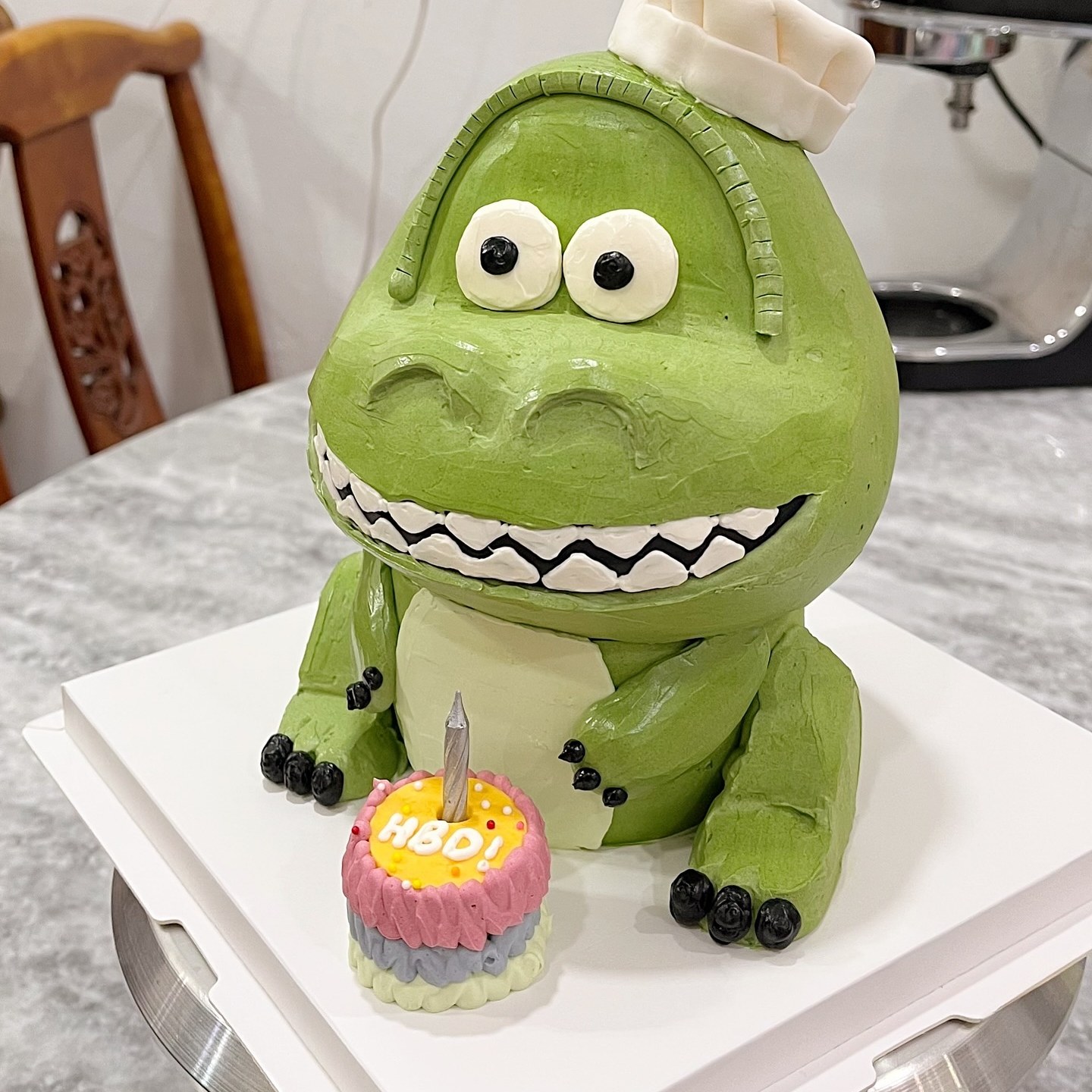 Green dinosaur character cake wearing a chef hat, with a mini birthday cake