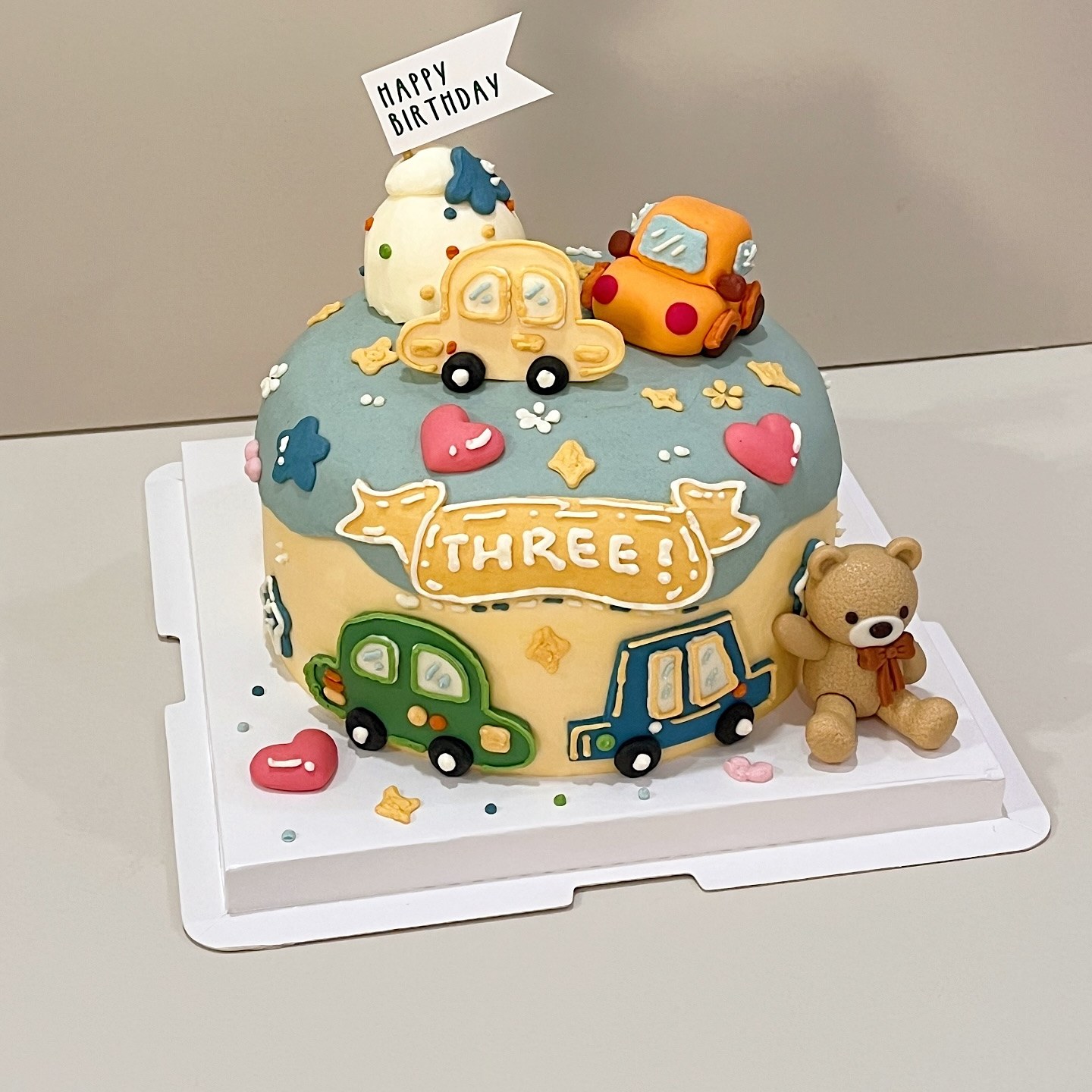 Blue and cream cake with car and truck decorations and a teddy bear figurine